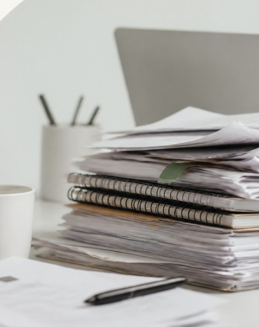 A tall stack of papers and notebooks on a cluttered desk